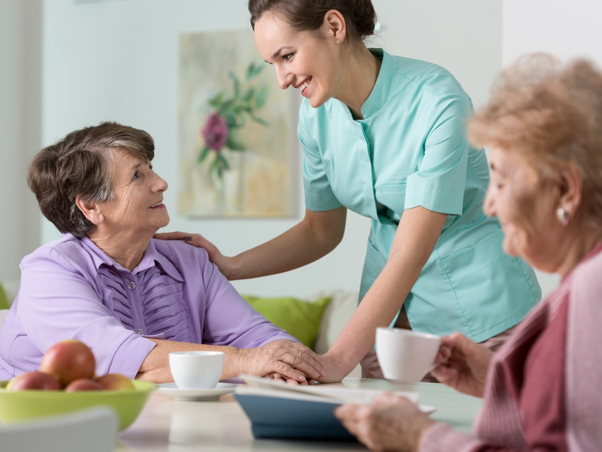 Zwei &auml;ltere Damen am Tisch mit einer Pflegerin, die die Hand auf die Schulter einer der Damen legt