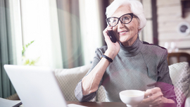 &Auml;ltere Dame mit wei&szlig;en Haaren sitzt vor einem Laptop und telefoniert