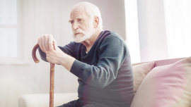Alter Mann sitzt auf einem Sessel und h&auml;lt sich an seinem Gehstock fest.