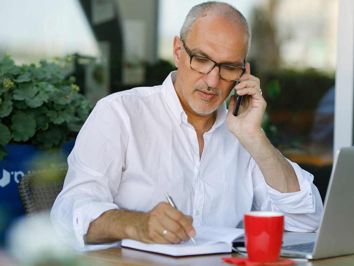 Mann mittleren Alters telefoniert und schreibt vor seinem aufgeklappten Laptopitzend