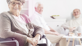 &Auml;ltere Frau im Rollstuhl lacht in die Kamera, im Hintergrund andere Senoioren