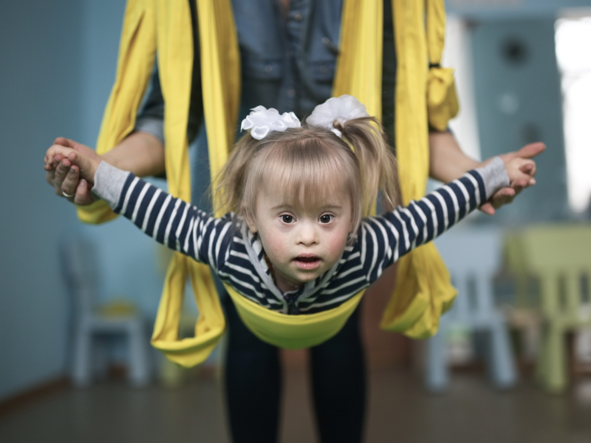Kleines M&auml;dchen, h&auml;ngt in einem gelben Tuch und fliegt, hinter ihm ein Erwachsener